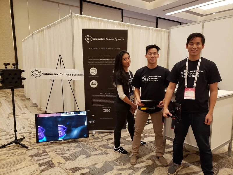 Three VCS team members posing beside the compact camera, branded banner and display.